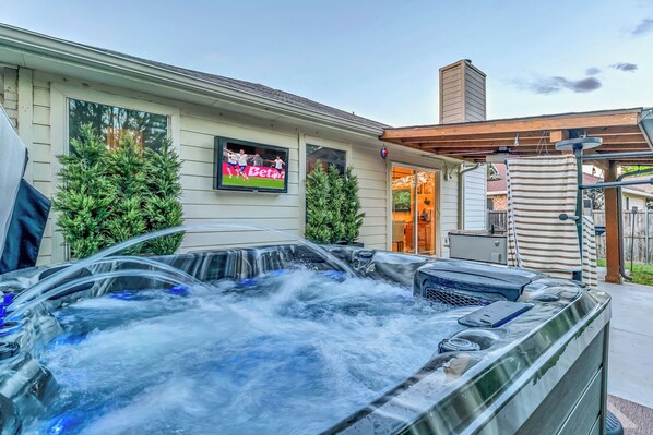A hot tub with a TV screen displaying a sports game, surrounded by greenery and a building with a chimney. - Backyard Paradise • Hot Tub • Fire Pit • 13 guests (McKinney)