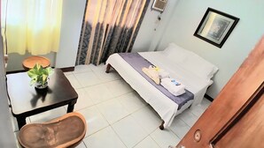 Una habitación de hotel con una cama, una mesita con una planta, una silla, un banco de madera y una puerta.