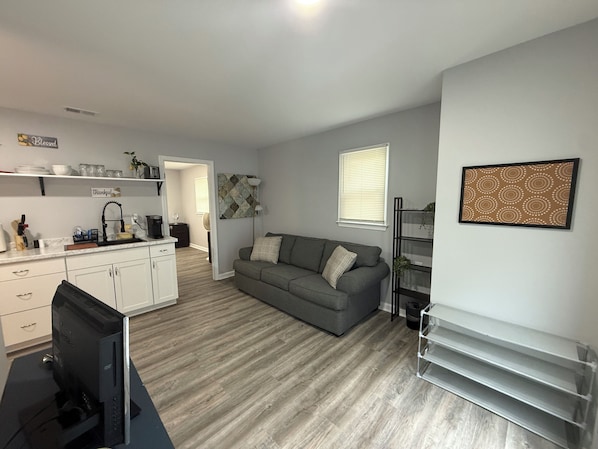 Un espacio habitable compacto con kitchenette, un sofá gris, un televisor de pantalla plana y una obra de arte enmarcada en la pared.