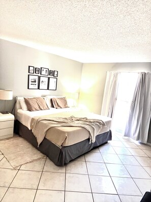 A bedroom with a bed, a nightstand, a lamp, a wall with framed pictures, and a window with curtains. - Beach Apartment Sunny Isles Steps from the Beach (Sunny Isles Beach)