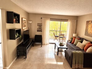 A living room with a sofa, TV, and a balcony with outdoor furniture. - Sunny Isles Beachfront Condo, Just Steps from the Beach (Sunny Isles Beach)