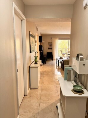 A hallway with a white door, a small white console table with a green bowl, and a window with curtains. - Sunny Isles Beachfront Condo, Just Steps from the Beach (Sunny Isles Beach)