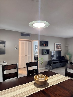 A dining area with a wooden table, chairs, a central plant, a television, and a ceiling light fixture. - Beach Apartment Sunny Isles Steps from the Beach (Sunny Isles Beach)