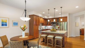 A modern kitchen with wooden cabinets, a central island with bar stools, pendant lighting, and a dining area with wicker chairs. - Pili Mai 12K Spacious 3BR with Central AC (Koloa)