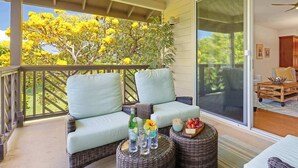A covered balcony with wicker furniture, a glass table with drinks, and a view of yellow flowering trees. - Pili Mai 12K Spacious 3BR with Central AC (Koloa)