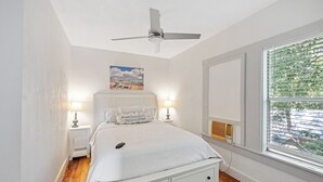 Une chambre à coucher comprenant un lit, des lampes de chevet, un ventilateur de plafond, une fenêtre avec des stores et un radiateur fixé au mur.