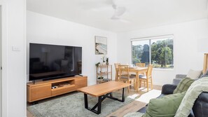 Ein modernes Wohnzimmer mit Flachbildfernseher, Holzmöbeln, einem Esstisch und einem großen Fenster mit Blick auf Grünflächen.