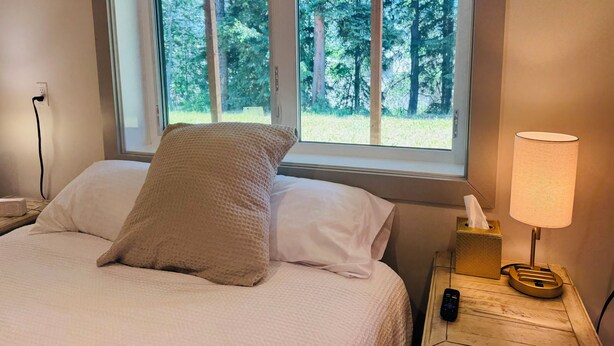 A bed with a textured pillow, a bedside lamp, and a window with a view of trees. - Modern, Clean and Cozy 2 Bedroom Duplex in Quiet West Fairbanks (Fairbanks)