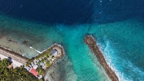 Vista aérea de una propiedad costera con un rompeolas rocoso, aguas turquesas cristalinas y un pequeño edificio con techo rojo.