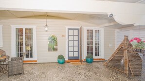 Een overdekte veranda met een wit plafond, twee deuren, een rieten stoel en een hanglamp.