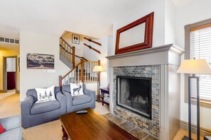 A living room with a fireplace, a sofa with decorative pillows, a wooden coffee table, and a staircase. - Charming Family Home: Summer Pool, Minutes to BM! (The Blue Mountains)