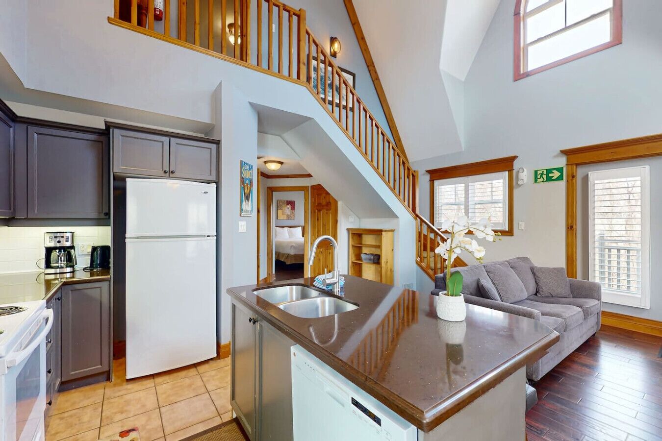 A kitchen with a refrigerator, stove, and coffee maker; a dining area with a sofa and a dining table; a staircase leading to an upper level; and a window with blinds.