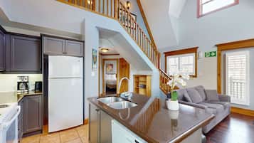 A kitchen with a refrigerator, stove, and coffee maker; a dining area with a sofa and a dining table; a staircase leading to an upper level; and a window with blinds.