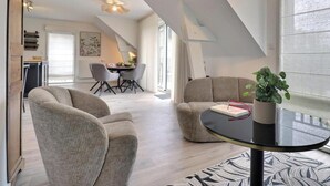 Ein modernes Wohnzimmer mit einer Couch, einem Couchtisch und einem Topf mit Pflanze. Im Hintergrund befindet sich ein Essbereich mit Tisch und Stühlen.