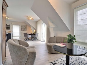 A modern living room with a sofa, a coffee table, and a potted plant. There is a dining area with a table and chairs in the background. - Le Clos de la Vicomté (DINARD)