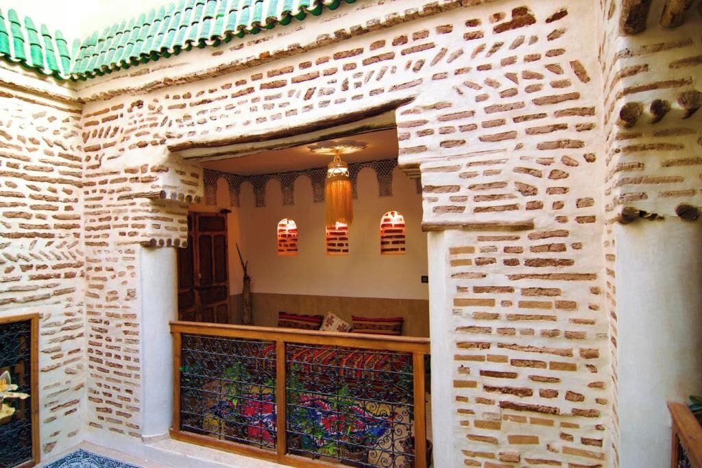 Una habitación con pared de ladrillo, ventana con celosías metálicas decorativas y techo de tejas verdes.