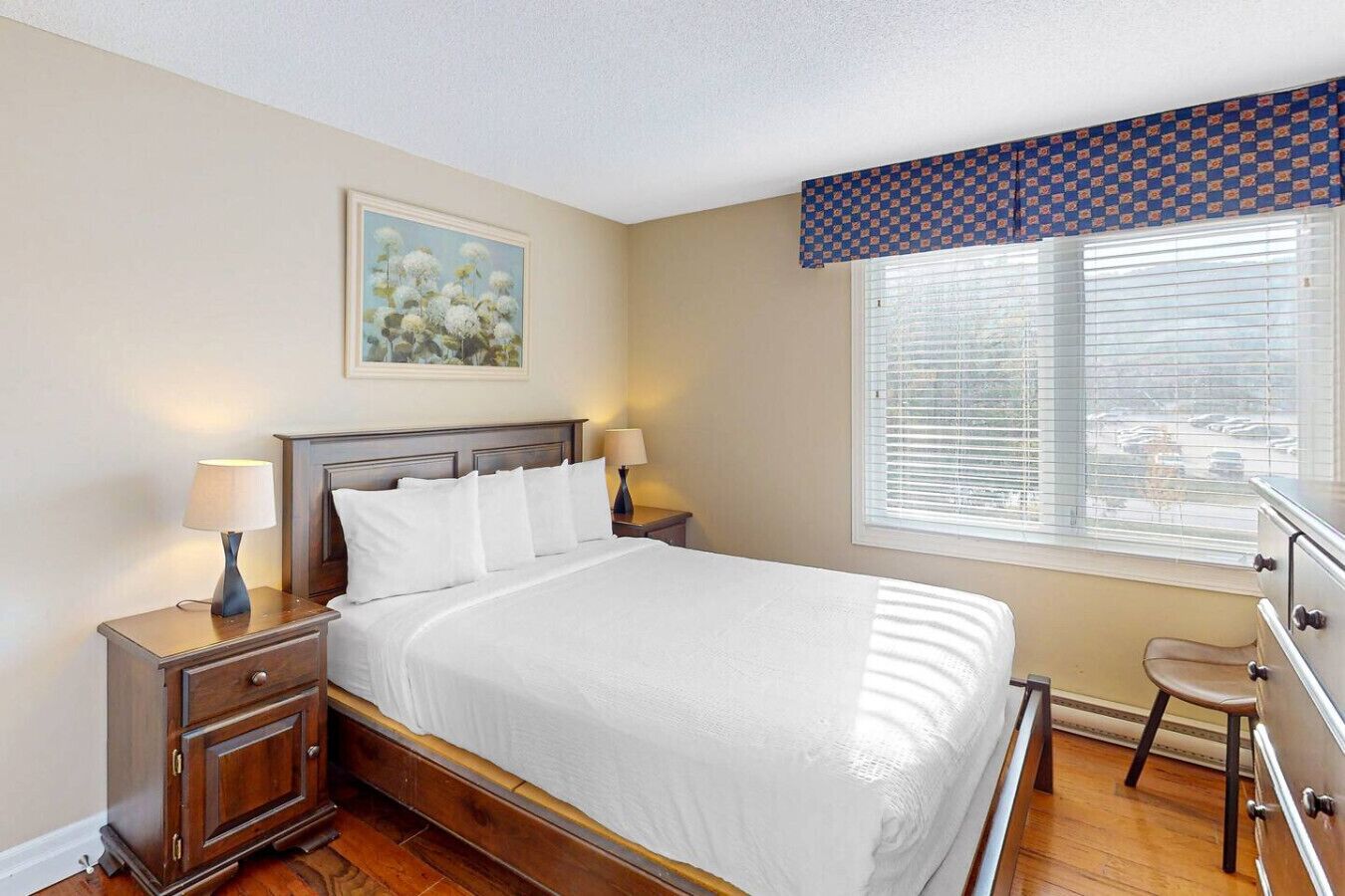 A bedroom with a bed, two bedside tables, a nightstand, a chair, a window with blinds, and a painting on the wall.