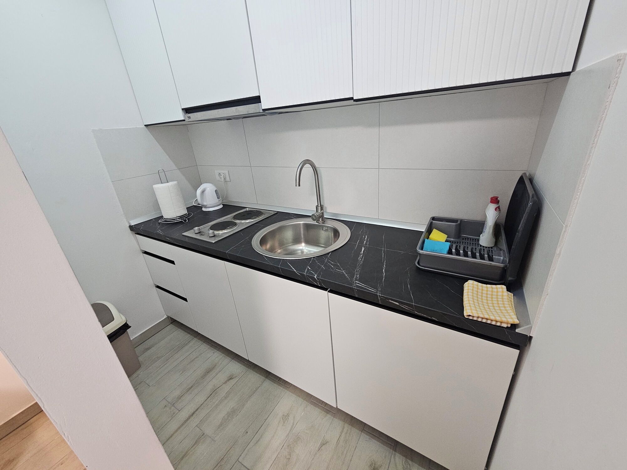 Een moderne keuken met witte kastjes, een roestvrijstalen spoelbak, een zwart aanrecht en een klein rekje voor afwasbenodigdheden.