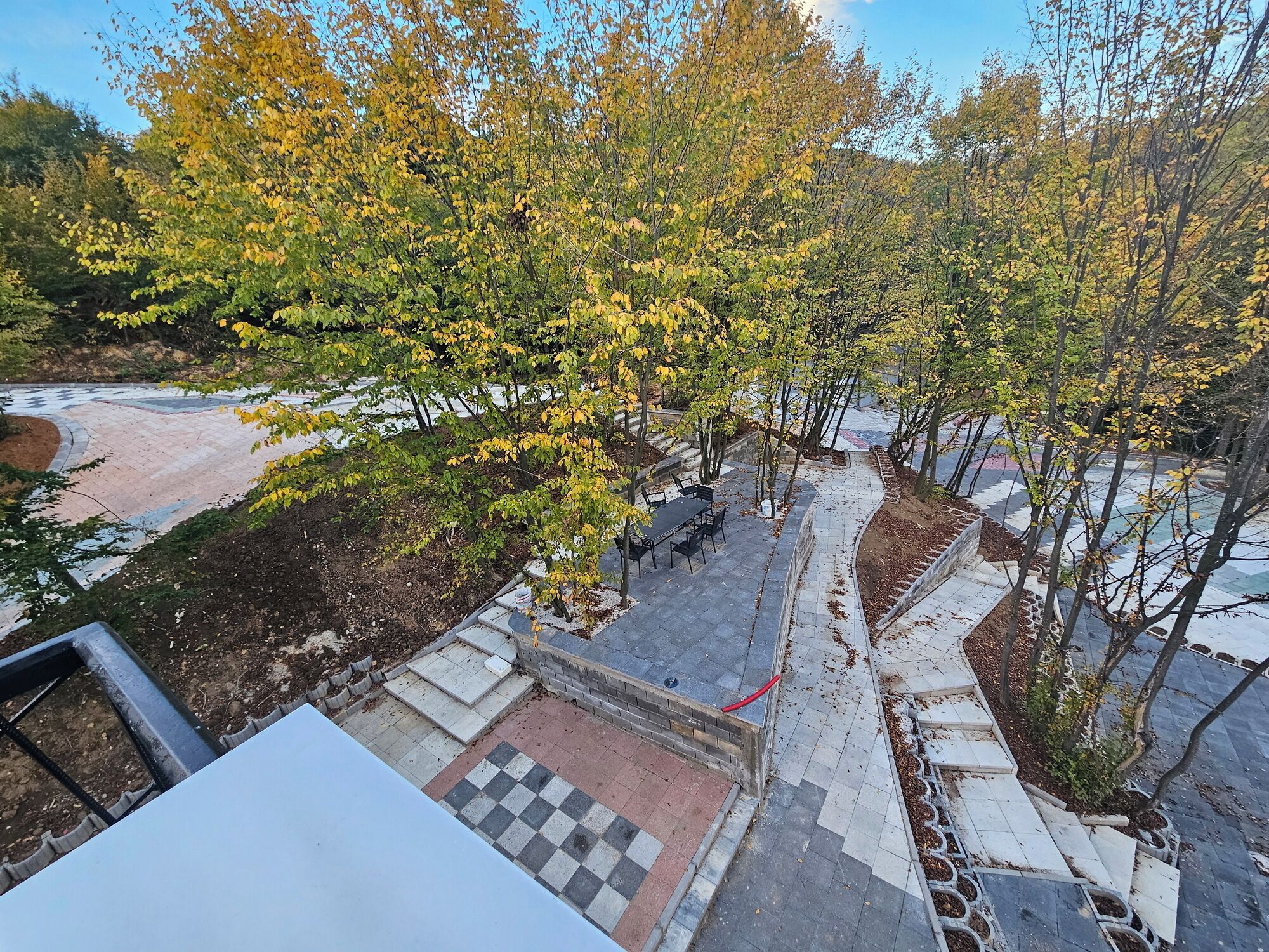 A paved outdoor area with a checkered pattern, a table with chairs, and trees with yellow leaves.