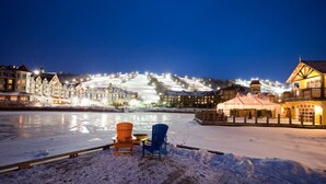 Ein Hotel am Wasserufer mit einer schneebedeckten Terrasse, zwei Adirondack-Stühlen und einem Gebäude mit schneebedecktem Dach im Hintergrund.