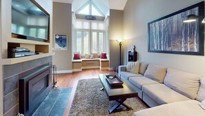 A living room with a fireplace, a sofa, a coffee table, a TV, and a window with a view of the outdoors. - Renovated 3BDR Family Condo | Slope-Side at Blue! (The Blue Mountains)
