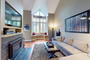A living room with a fireplace, a sofa, a coffee table, a TV, and a window with a view of the outdoors. - Renovated 3BDR Family Condo | Slope-Side at Blue! (The Blue Mountains)