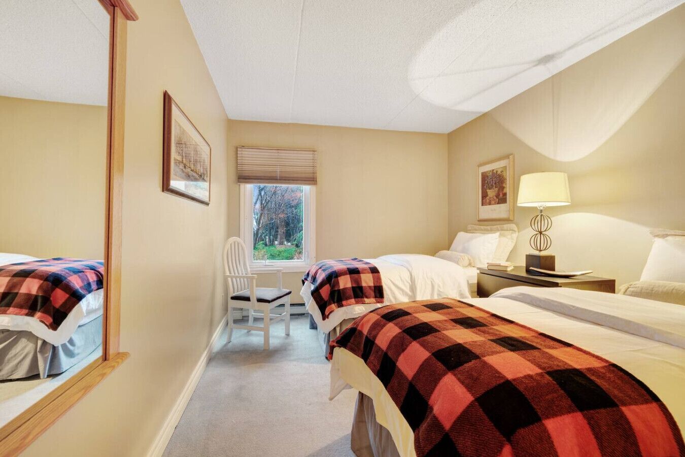 Ein Hotelzimmer mit zwei Betten, einem Schreibtisch, einem Stuhl, einem Fenster mit Jalousien, einer Lampe und einem gerahmten Bild an der Wand.