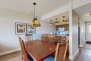 A dining area with a wooden table and chairs, pendant lights, framed artwork on the wall, and a kitchen in the background. - Golf-Side Retreat: Pond Views w/ Summer Pool (The Blue Mountains)