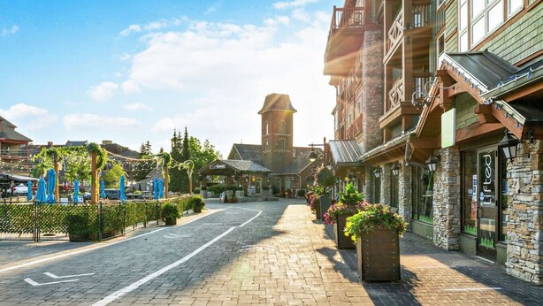 Una strada acciottolata fiancheggiata da edifici in pietra, una torre campanaria e aree con tavoli all'aperto coperte da ombrelloni.