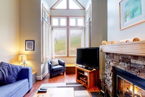 Uma sala de estar com lareira, televisão, sofá e janela com vista para o exterior.