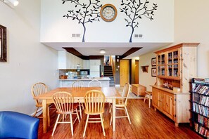Uma sala de jantar com uma mesa e cadeiras de madeira, uma cozinha ao fundo e uma estante de livros.