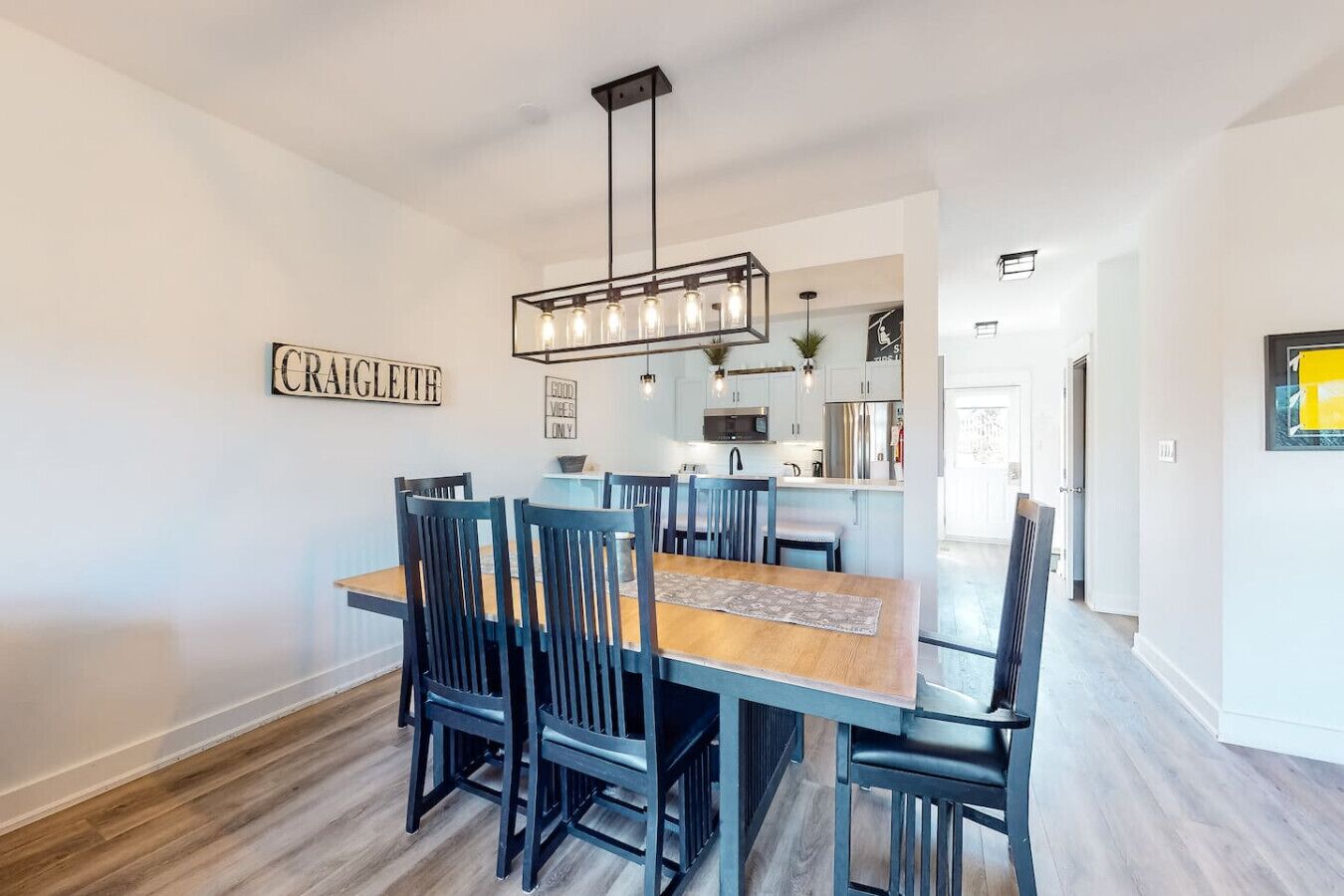 A dining area with a wooden table and chairs, a hanging light fixture, and a kitchen in the background.