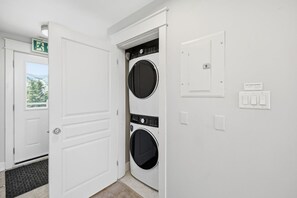 A laundry room with two stacked washing machines, a door with a doormat, and a wall-mounted light switch. - Rivergrass Sanctuary: Hot Tub & Summer Pool (The Blue Mountains)
