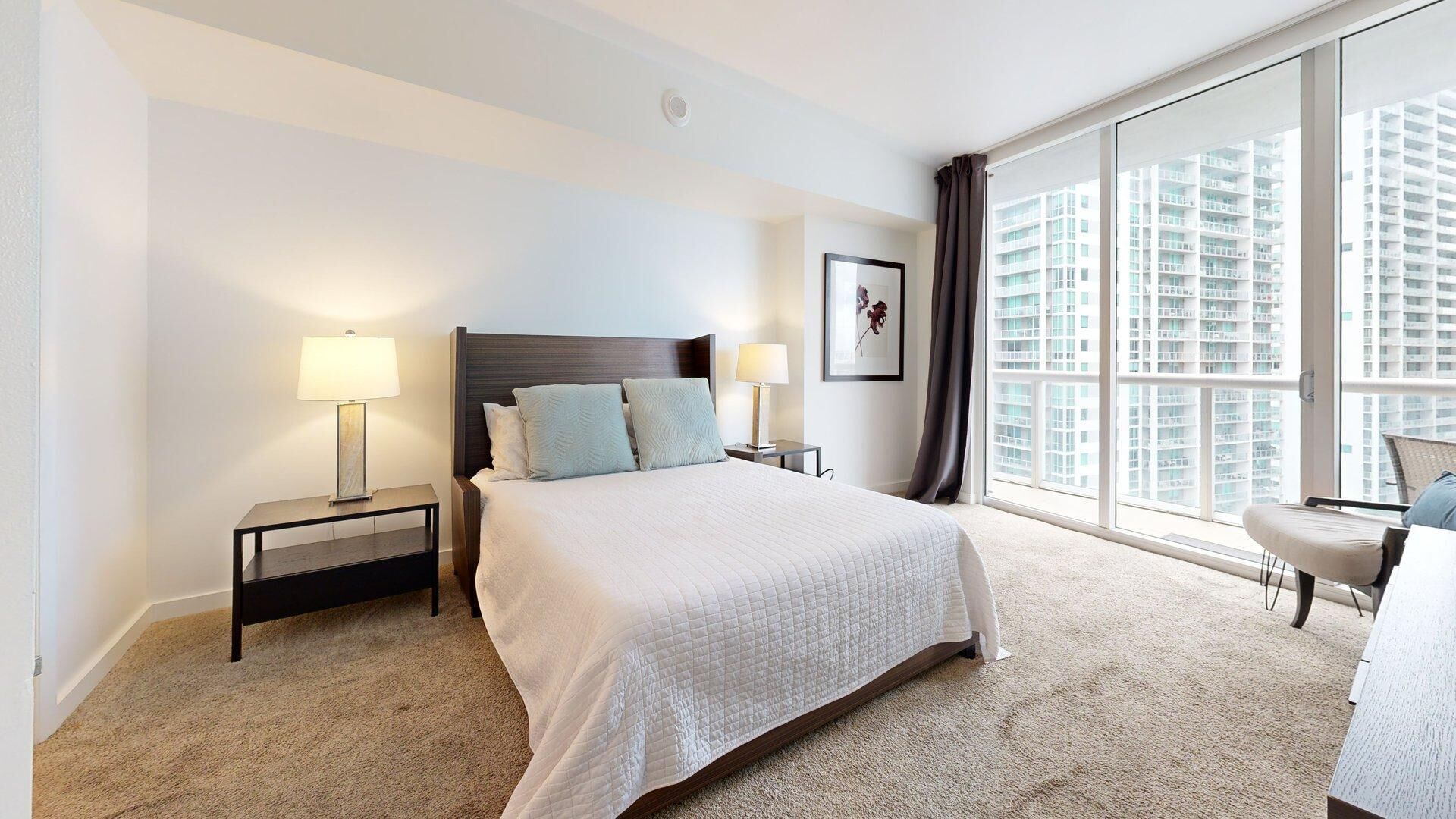 A hotel room with a large bed, two bedside tables with lamps, a chair, a large window with a city view, and a framed picture on the wall.