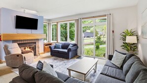 A modern living room with a fireplace, a flat-screen TV, and a large window offering a view of the outdoors. - 3 Bedroom Home | Steps to Blue Mtn Village (The Blue Mountains)