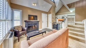 A living room with a fireplace, a sofa, a television, and a staircase leading to a kitchen. - Spacious North Base Condo - Pool, Hot Tub, Tennis (The Blue Mountains)