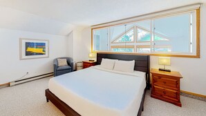 Ein Schlafzimmer mit einem großen Bett, einem Sessel, einem Nachttisch, einer Lampe und einem Fenster mit Blick auf Bäume.