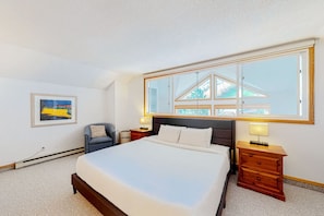 Ein Schlafzimmer mit einem großen Bett, einem Sessel, einem Nachttisch, einer Lampe und einem Fenster mit Blick auf Bäume.