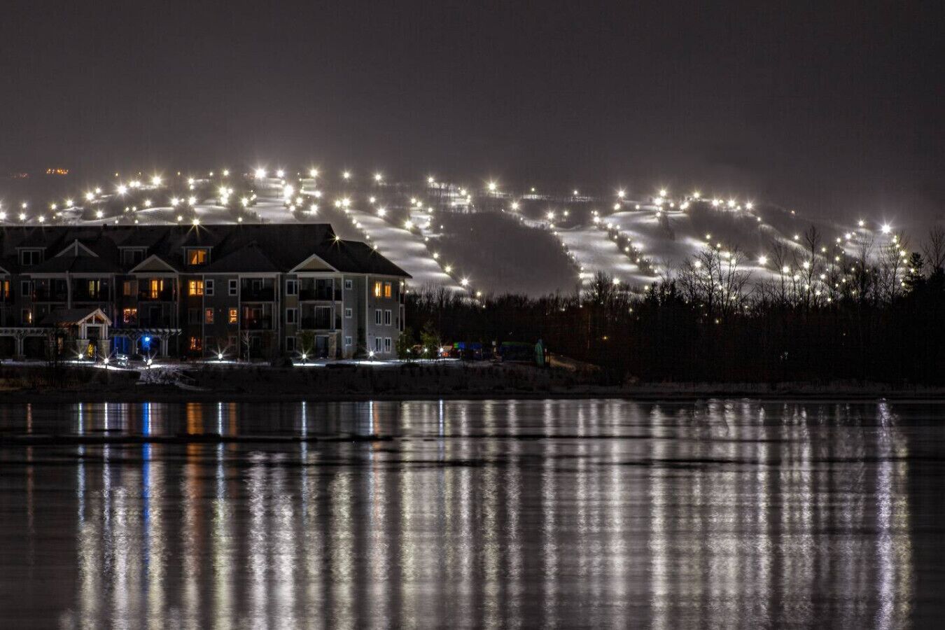 Une propriété située au bord de l’eau, avec une rangée de bâtiments illuminés, une surface d’eau qui reflète la lumière et une colline lointaine illuminée.
