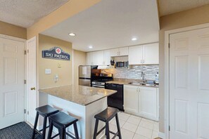 A kitchen with white cabinets, a granite countertop, a stainless steel refrigerator, a microwave, and a dishwasher. There are two black stools at the island. - 1BDR North Creek with Pool, Hot Tub, Tennis! (The Blue Mountains)