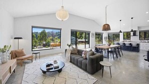 A spacious living area with a sofa, coffee table, and dining table, large windows with a view of greenery, and a modern kitchen.