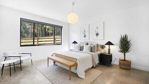 A bedroom with a large window, a bed, a bench, a bedside table, and a lamp.