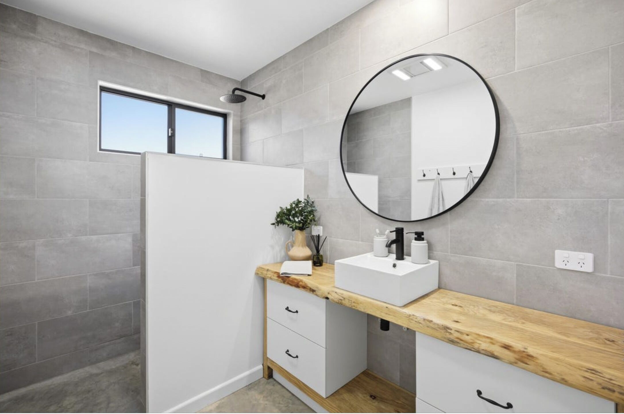 A modern bathroom with grey tiles, a wooden countertop, a white sink, a round mirror, and a window.