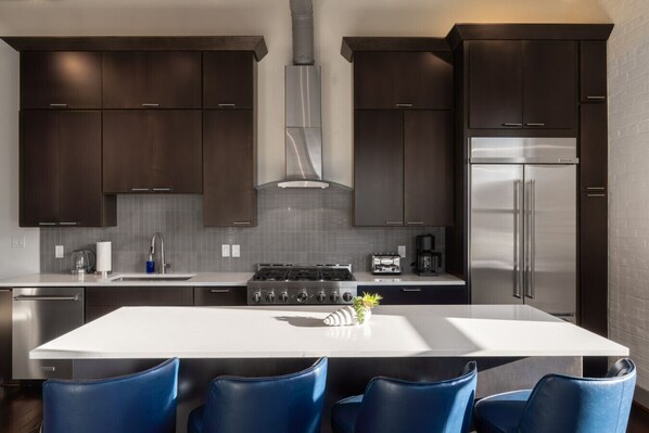 A modern kitchen with dark wood cabinets, a stainless steel refrigerator, a gas stove, and blue chairs around a central island. - Luxury 3BR on TQL Stadium & Washington Park (Cincinnati)