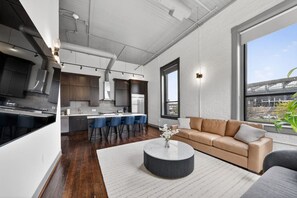 A modern kitchen with dark cabinets, a central island with bar stools, a large window, and a beige sofa in a living area. - Luxury 3BR on TQL Stadium & Washington Park (Cincinnati)