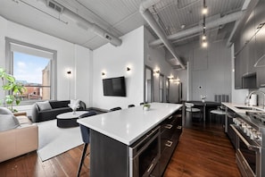 A modern kitchen with a central island, a dining area with a sofa and chairs, and a large window providing a view of the city. - Luxury 3BR on TQL Stadium & Washington Park (Cincinnati)