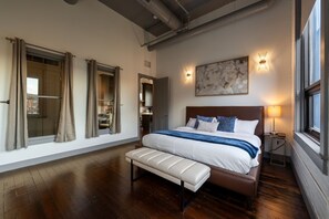 A bedroom with a large bed, a bench, a nightstand, a wall-mounted lamp, and a window with curtains. - Luxury 3BR on TQL Stadium & Washington Park (Cincinnati)