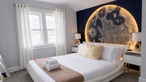 Een hotelkamer met een groot bed, witte gordijnen, een muurschildering met een maanmotief en bedlampjes.