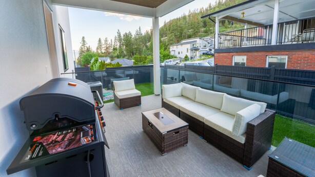 En uteplats med en grill, möbler i rotting och ett glasräcke med utsikt över ett bostadsområde.