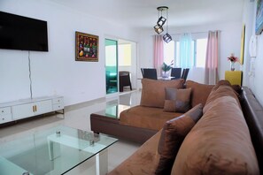 A modern living room with a glass coffee table, a brown sofa, a flat-screen TV mounted on the wall, and a dining area with a table and chairs. - Malecon dream, Splendid air-conditioned apartment with pool and parking (Santo Domingo Este)